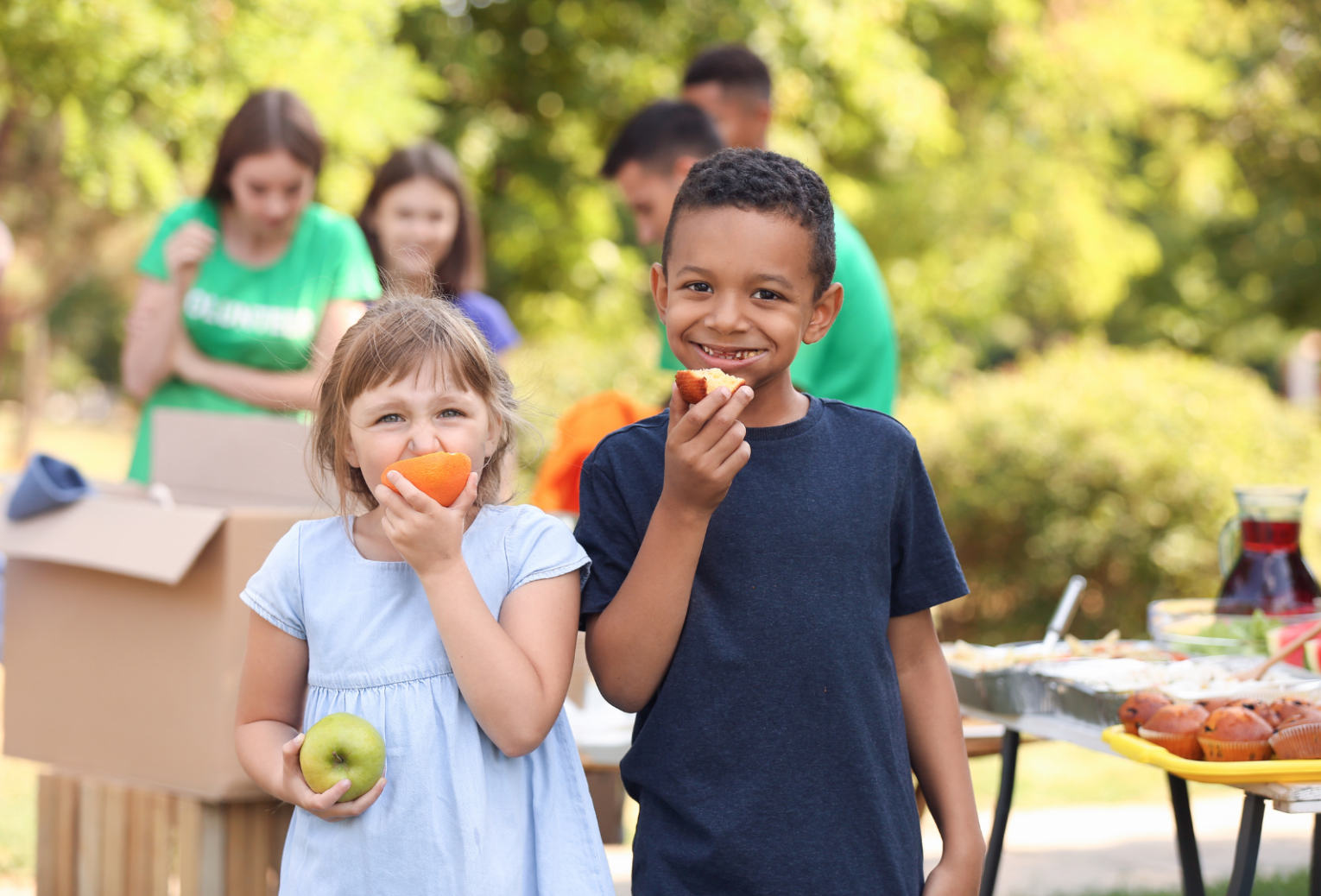 A small white girl and a small Black boy eating apples at a cookout with adults in the background